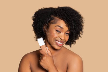 Schöne schwarze Frau mit natürlichen Locken hält das Fundament an ihre Wange, ihr strahlendes Lächeln spiegelt die Freude wider, ihr Make-up zu perfektionieren