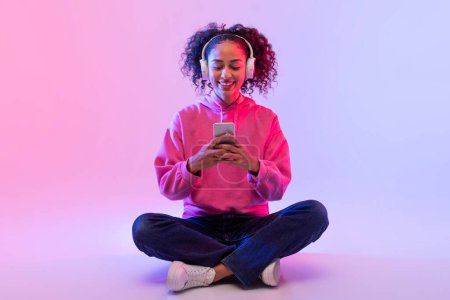 Foto de Mujer joven y relajada con capucha rosa usando un teléfono inteligente mientras está sentada con las piernas cruzadas en el suelo - Imagen libre de derechos