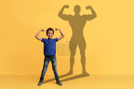 Téléchargez les photos : Un garçon joyeux dans une chemise bleue fléchit ses muscles avec un grand sourire, jetant une silhouette de super-héros forte sur un fond jaune, illustrant la confiance et les rêves des jeunes - en image libre de droit