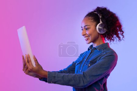 Foto de Mujer sonriente en chaqueta de mezclilla y auriculares sostiene una tableta brillante, color retroiluminado - Imagen libre de derechos