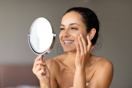 Photo for Cheerful young woman smiles while applying moisturizer holding mirror, promoting beauty and facial skincare, closeup in modern bedroom interior. Concept of rejuvenation for smooth healthy skin - Royalty Free Image