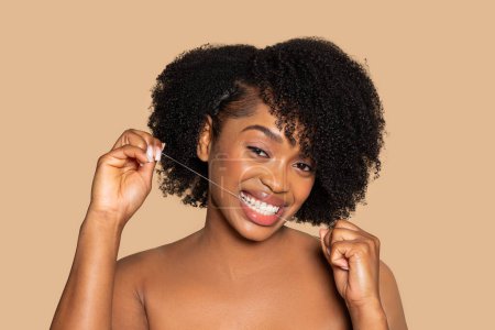 Photo for African american woman with curly hair joyfully flossing her teeth, showcasing wide, engaging smile against warm beige backdrop, promoting dental health - Royalty Free Image