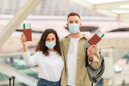 Photo for Young European Couple Wearing Medical Masks In Airport Holding Passports And Tickets, Happy Millennial Man And Woman Enjoying Safe Travels, Ready For Flight, Looking At Camera, Free Space - Royalty Free Image