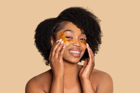 Photo for Radiant young black woman with natural afro hairstyle enjoys pampering session with gold hydrogel eye patches on soft beige background, self-care concept - Royalty Free Image