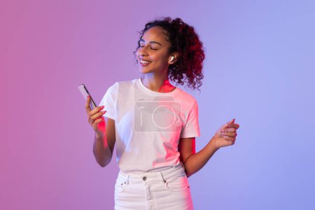 In a vibrant neon background, a woman with curly hair holds a smartphone while making a hand gesture