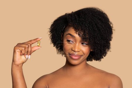 Playful young black woman with natural curly hair showing golden nutritional supplement capsule and smile on creamy background