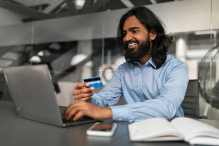 Man in a blue shirt holds a credit card and smiles while shopping online at his office workspace