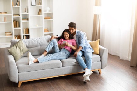 Téléchargez les photos : Couple hindou adorant blotti sur un canapé gris, regardant confortablement le smartphone ensemble, dans un environnement confortable avec un éclairage chaud, pleine longueur - en image libre de droit