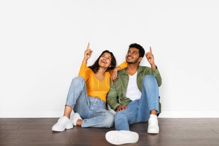 Photo for Joyful Indian couple in casual clothes sitting on wooden floor, smiling and pointing upwards at free space with white background enhancing their casual style - Royalty Free Image