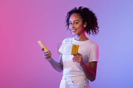 Téléchargez les photos : Une jeune femme joyeuse regarde un smartphone tout en tenant une carte de crédit, symbolisant le commerce mobile - en image libre de droit
