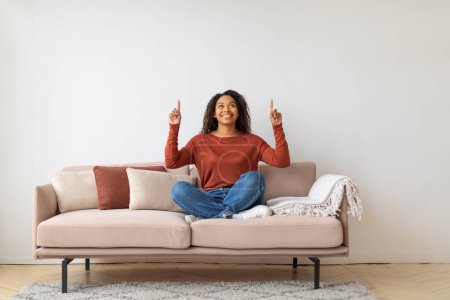 Photo for Cheerful young black woman sitting cross-legged on sofa, smiling and pointing upwards, happy african american female suggesting idea or concept while relaxing in bright living room, copy space - Royalty Free Image