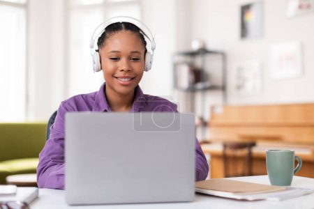 Téléchargez les photos : Étudiante afro-américaine engagée avec des tresses et des écouteurs souriants, tout en utilisant un ordinateur portable pour une étude en ligne dans un cadre confortable à la maison avec une tasse de thé chaude. - en image libre de droit