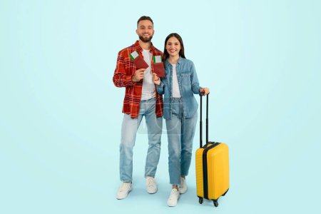 Téléchargez les photos : Joyeux couple de voyageurs caucasiens qui attendent avec impatience des vacances de rêve, debout avec des billets de voyage et une valise, souriant alors qu'ils planifient une escapade romantique, posant sur fond bleu. Longueur totale - en image libre de droit