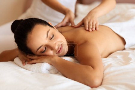 Téléchargez les photos : Soin du corps. Jeune femme détendue profitant d'un massage et d'une touche apaisante de masseuses mains sur le dos enveloppées dans une serviette blanche sur le lit intérieur. Spa, procédures beauté - en image libre de droit