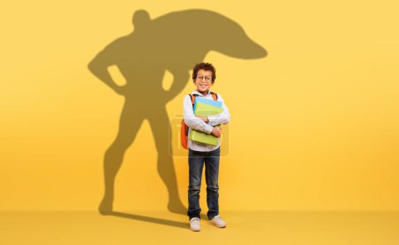Téléchargez les photos : Un jeune garçon studieux aux cheveux bouclés et aux lunettes tient fermement ses livres scolaires, fièrement debout devant un fond jaune avec une ombre de super-héros signifiant force et ambition - en image libre de droit
