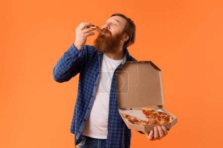 Photo for Caucasian redhaired bearded guy in casual enjoys cheat meal with pizza, holding delivery box and savoring tasty slice against orange studio background. Concept of overeating and junk food - Royalty Free Image