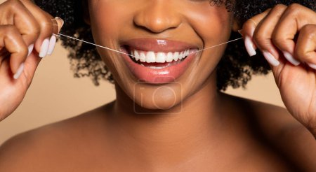 Nahaufnahme eines strahlenden Lächelns, als schwarze Frau mit schönem lockigem Haar ihre Zähne gekonnt mit Zahnseide pflegt und auf neutralem Hintergrund Mundhygiene fördert
