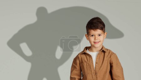 Téléchargez les photos : Un jeune garçon heureux dans une veste brune sourit vivement, debout devant une toile de fond grise avec une ombre suggérant qu'il porte un chapeau et un manteau détectives, faisant allusion à l'aventure et à la curiosité - en image libre de droit