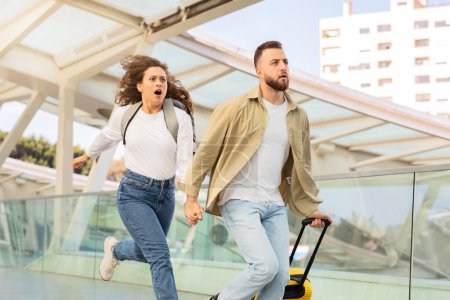 Stressed young couple running with luggage in airport terminal, late for their flight, in motion shot of emotional man and woman in rush, holding hands while going to flight gate, free space