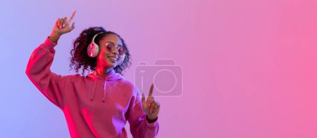 Foto de Mujer joven feliz en sudadera con capucha rosa y gafas de sol bailando con auriculares, gestos y sonrisas, en neón rosa y azul degradado fondo, panorama, espacio libre - Imagen libre de derechos