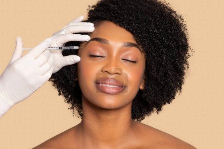 Foto de Mujer negra pacífica con los ojos cerrados experimenta inyección facial, destacando su confianza en los tratamientos estéticos para la perfección de la piel en tono beige - Imagen libre de derechos