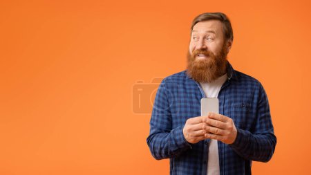 Téléchargez les photos : Homme barbu aux cheveux roux souriant tenant son téléphone portable et regardant de côté l'espace vide, la maquette pour l'offre de publicité, les textos de gars et l'utilisation de l'application sur l'appareil, sur fond orange. Panorama - en image libre de droit