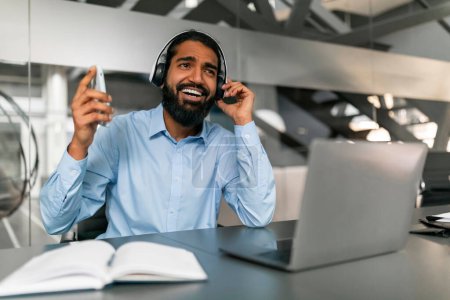 Professionnel joyeux utilisant un casque et tenant un smartphone dans un cadre de bureau moderne, indiquant une communication à distance