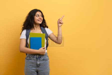 Curious female student pointing upwards, posing with blue notebooks, indicating interest and ideas