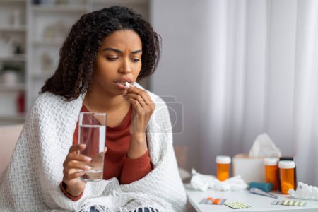 Foto de Mujer negra joven sintiéndose mal en casa, tomando píldora con vaso de agua, mujer afroamericana enferma usando medicina mientras está sentada envuelta en una manta en el sofá en la sala de estar, espacio para copiar - Imagen libre de derechos