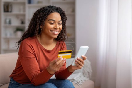 Foto de Mujer negra sonriente en casa con tarjeta de crédito y teléfono inteligente, joven afroamericano feliz compras en línea o haciendo pagos móviles mientras se relaja en la acogedora sala de estar, espacio para copiar - Imagen libre de derechos