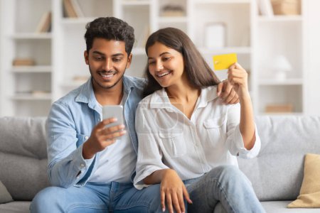 Téléchargez les photos : Jeunes conjoints indiens détendus effectuant des achats en ligne à la maison, tenant carte de crédit et smartphone tout en choisissant des produits dans un salon lumineux et confortable - en image libre de droit
