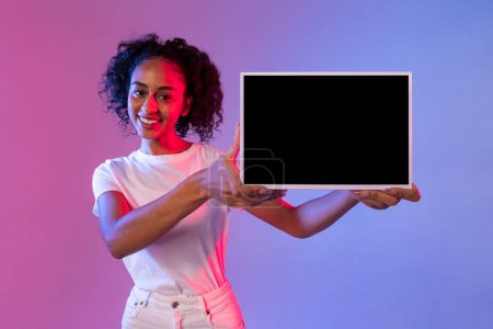 Photo for Smiling black lady presenting digital tablet with black screen, ideal for app display, mockup, ad, against blended pink and blue gradient neon background - Royalty Free Image