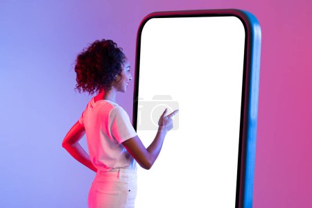 Téléchargez les photos : Une dame en blanc se tient face à un gigantesque écran de smartphone vide, représentant les technologies à grande échelle dans nos vies - en image libre de droit