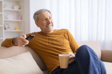 A relaxed senior male lounges on a couch holding a TV remote and a mug, looking away thoughtfully as if watching TV