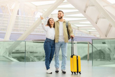 Photo for Happy young couple enjoying their time at the airport while waiting flight, cheerful millennial man and woman standing with yellow luggage at terminal hall, woman pointing away and smiling - Royalty Free Image