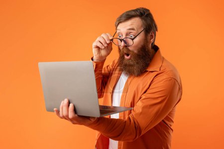 Photo for Amazed freelancer guy with red hair and beard looking at laptop computer, touching his glasses in shock, standing with notebook while working online and websurfing on orange studio background - Royalty Free Image