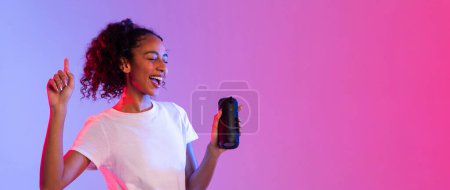 Foto de Enérgica dama negra en blanco bailando y sonriendo, sosteniendo altavoz portátil con expresión alegre, frente a vibrante fondo de neón rosa de doble tono, panorama, espacio libre - Imagen libre de derechos