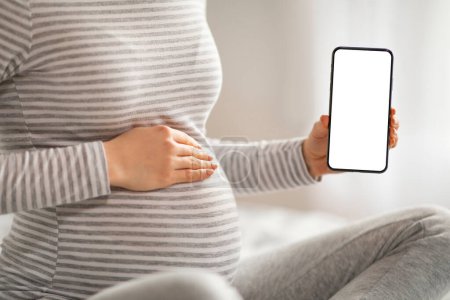 Schwangerschaft App. Unerkennbare Schwangere demonstriert Smartphone mit leerem weißen Bildschirm, empfiehlt neue mobile Anwendung für werdende Frauen, Attrappe mit Kopierraum