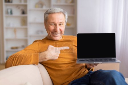 Foto de Anciano se sienta en un sofá, apuntando a la pantalla en blanco de un ordenador portátil, perfecto para la publicidad o mensajes educativos - Imagen libre de derechos