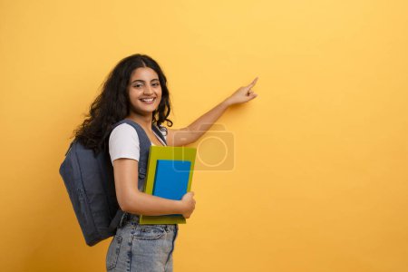 Téléchargez les photos : Étudiant souriant avec sac à dos pointant latéralement sur fond jaune, évoquant le choix ou la direction - en image libre de droit