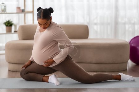 Téléchargez les photos : Concept de grossesse active. Souriant dame noire future maman étirant ses jambes, faire son entraînement de yoga matinal sur tapis à la maison gymnase. Jolie femme enceinte faisant du fitness domestique, faisant de l'exercice à l'intérieur - en image libre de droit