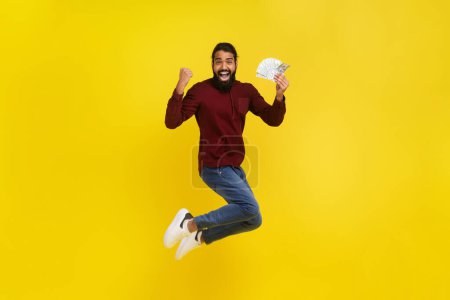 Photo for Happy indian guy winner jumping in the air with money in his hand, gesturing raising hand up, celebrating success, won lottery or got cashback, isolated on yellow studio background, full length - Royalty Free Image