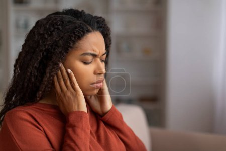 Stressed young black woman holding her ears and grimacing in pain, sick african american female feeling unwell at home, indicating ear infection or severe earache, sitting on couch in living room