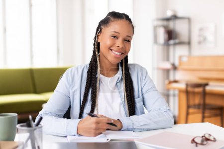 Photo for Cheerful african american female student with braids smiles as she writes in notebook, sitting at table with her laptop, exuding relaxed study vibe at home - Royalty Free Image