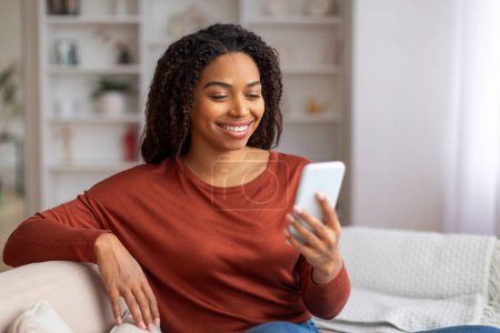 Photo for Beautiful Young African American Woman Relaxing With Smartphone At Home, Happy Black Female Sitting On Couch And Holding Mobile Phone, Reading Message Or Browsing New App, Free Space - Royalty Free Image