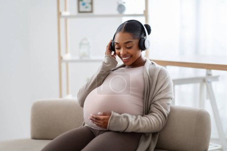 Téléchargez les photos : Jeune femme enceinte afro-américaine souriante écoutant de la musique, assise sur un canapé à la maison, embrassant son gros ventre, utilisant un casque sans fil, profitant de ses chansons préférées, copiant de l'espace - en image libre de droit