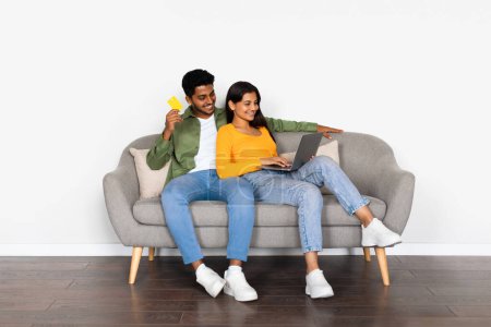 Interesantes indisches Paar beim Online-Shopping, Kreditkarte und Laptop in der Hand, während es sich auf einer grauen Couch bequem macht, in minimalistischem Raum