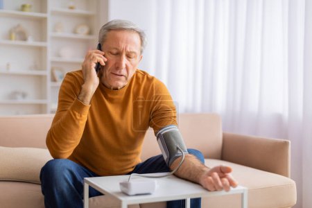 Foto de Un anciano preocupado está sentado en un sofá usando un monitor de presión arterial y hablando por teléfono en su casa. - Imagen libre de derechos