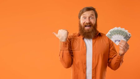 Téléchargez les photos : Homme barbu aux cheveux roux excité avec des points d'argent liquide à part exsudant joie et succès, affichant une poignée de billets en euros et un espace vide pour la publicité, sur fond de studio orange. Panorama - en image libre de droit
