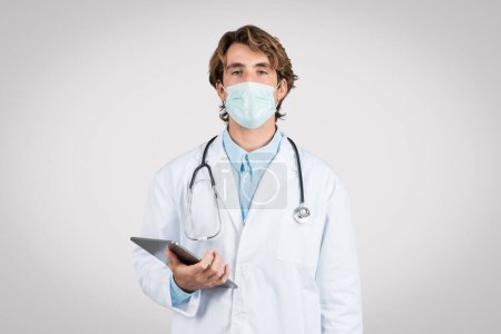 Foto de Médico masculino enfocado usando máscara quirúrgica y estetoscopio, usando tableta digital, significando salud y fusión de la tecnología, fondo gris - Imagen libre de derechos